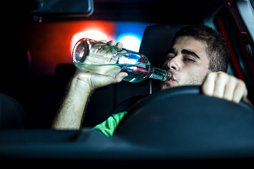 Police in pursuit of a reckless drunk driver at night. About 30 years old Caucasian man, drinking alcohol from the bottle.