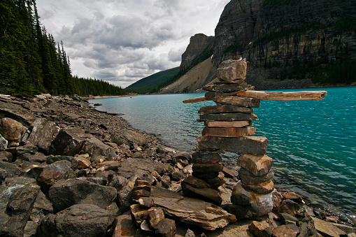 A stone Inukshuk stands on the shores of a lake in the Rocky Mountains in Canada. Inukshuks can be found throughout Canada, but are especially popular in the west and the north - from where they originate. This one was near a hiking trail in the Rocky Mountains, surrounded by beautiful mountains, green forests, and a stunning blue lake.