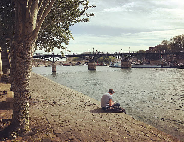 Paris, France - September 26, 2016: People relaxing on Seine riverbank, Paris. Young man sitting on embankment reading a book. 
