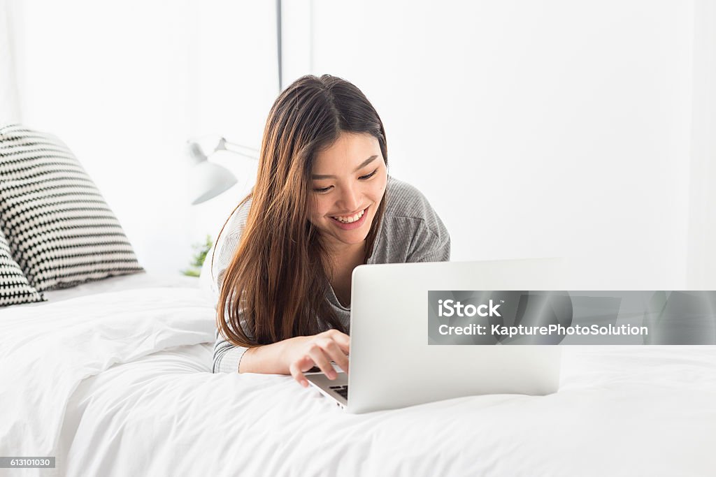 Asian girl laying on bed playing laptop Asian girl laying on bed playing laptop and smiling Laptop Stock Photo