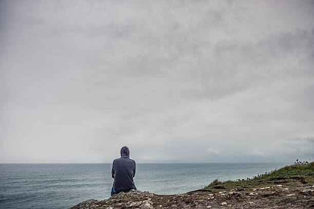 istock Man wearing hoodie sat on cliffs above water. 613130388