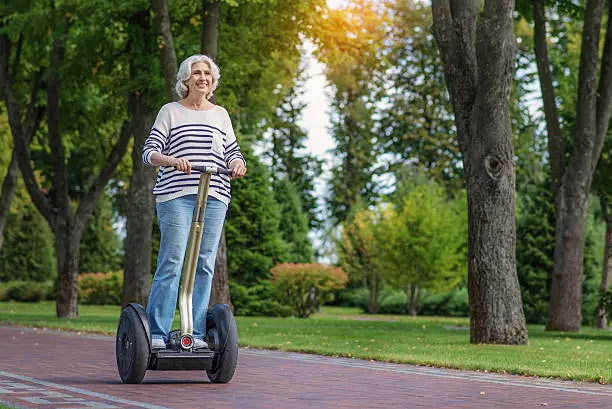 Joyful old woman is having fun in park. She is standing on Segway and laughing