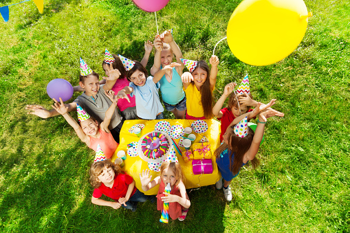 Top view of laughing kids standing around the table with birthday cake, waving hands and balloons, at the outdoor party