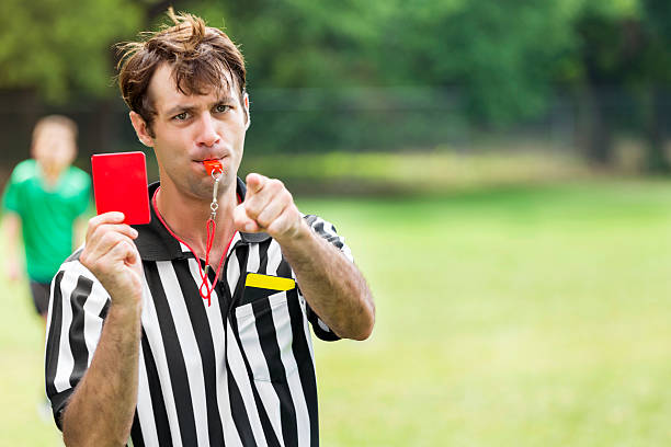Soccer referee points and holds up red card Serious mid adult Caucasian male referee looks at the camera and points while holding up a red card. he is also blowing a whistle. A young soccer player wearing a green jersey is in the background. Copy space is available. referee stock pictures, royalty-free photos & images