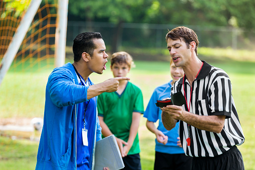 Mid adult Hispanic soccer coach yells at mid adult Caucasian referee and points at him over a bad call. The coach is wearing a blue hoodie and is holding a clipboard. The players in green jerseys and blue jerseys are watching and listening in the background.