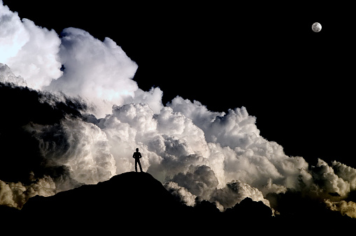 Standing alone on a barren mountain peak at high altitude, a man is dwarfed into insignificance and thrown into silhouette against the backdrop of a boiling Cumulonimbus storm cloud beneath a rising full moon.