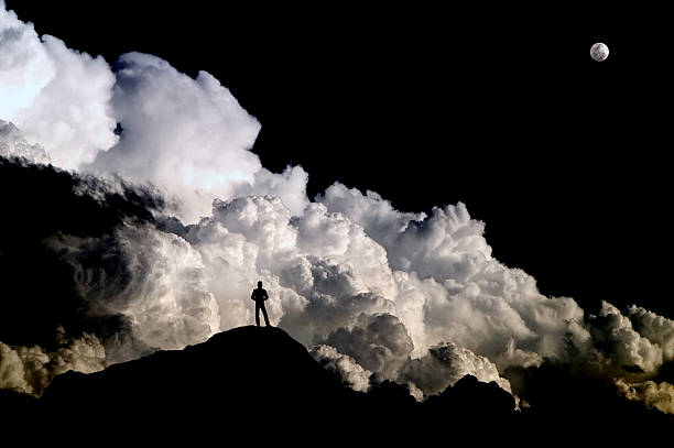Man standing on mountain silhouetted against turbulent storm clouds Standing alone on a barren mountain peak at high altitude, a man is dwarfed into insignificance and thrown into silhouette against the backdrop of a boiling Cumulonimbus storm cloud beneath a rising full moon. cumulonimbus photos stock pictures, royalty-free photos & images