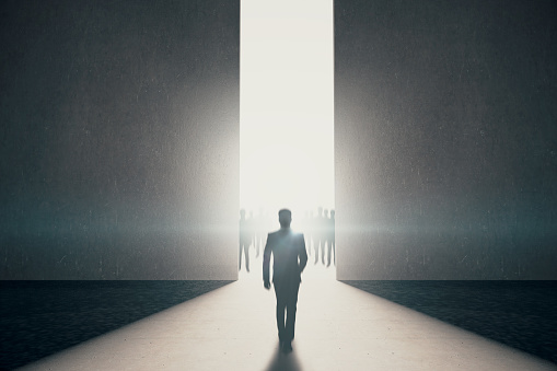 Back view of businessman walking towards abstract opening with bright light and people silhouettes in concrete interior. Success concept. 3D Rendering