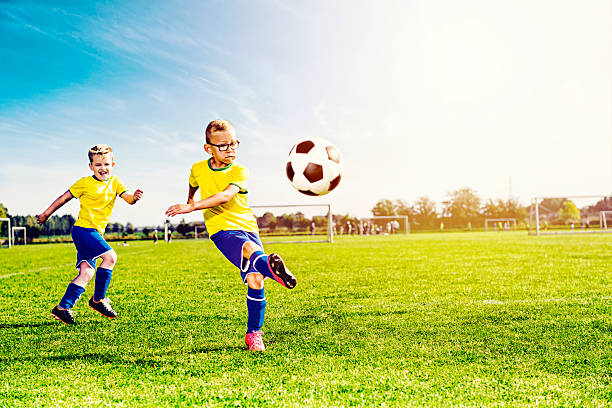 Teenage male soccer player kicks the ball during soccer game. The ball is in mid air. The boy is seen fromt he waist down. He is wearing a green jersey, black shorts, black socks and cleats. The opposing team is in blue jerseys. The opposing team and the boy's teammates are in the distance along with the soccer goal.