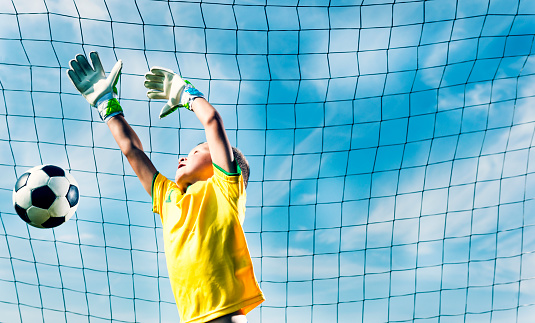 Boy around 6 years old wears sports gloves and goalkeeper outfit. He tries to block a shot and jumps high in the air.