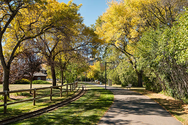 Horizontal stock photo of a walking path in Kinsmen Park in Saskatoon in autumn.  Colourful trees line both sides of the path which curves off into the distance.  The train tracks to the left of the image are for the children's train ride that is a popular attraction during the summer.  On the left in the background is the train barn where the train is stored when not in use.