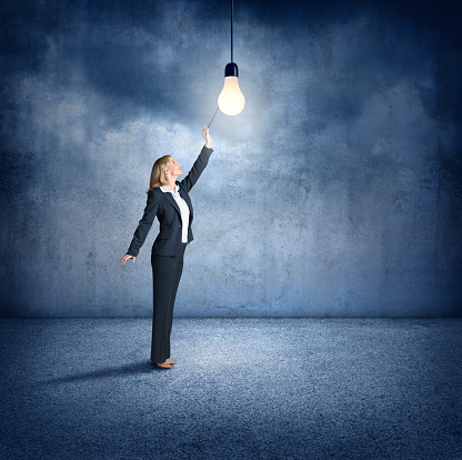 A businesswoman stands on her tiptoes as she reaches up to turn on a bare light bulb that is hanging above her.  She stands in a large cavernous room that is surrounded by concrete walls.