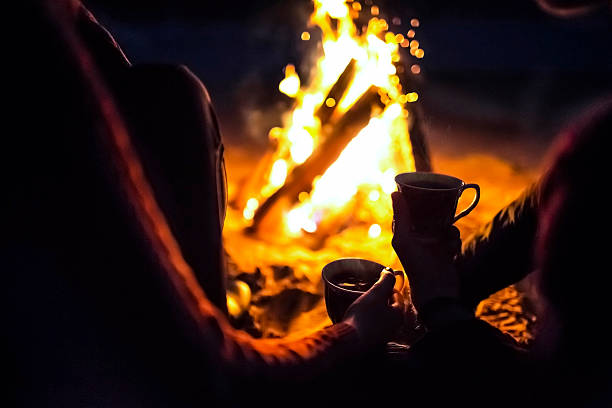 man and girl sit around campfire at night with cups stock photo