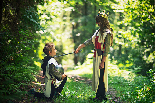 Young queen or teenage princess wearning knight's armour and a crown is knighting loyal knight with a sword. Green forest of a sunny summer day.