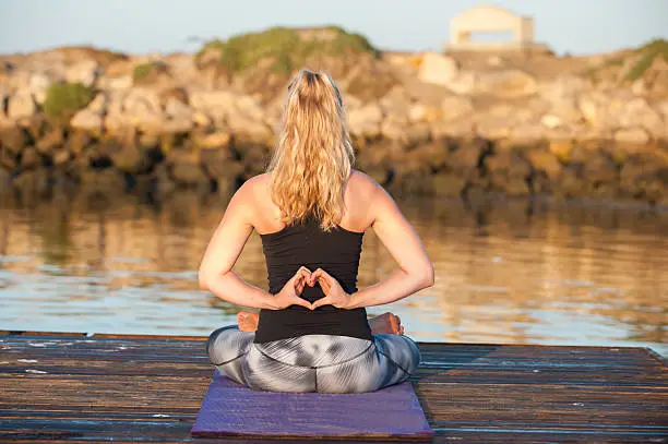 Athletic female seated on dock with hands behind back in heart shape while facing away.