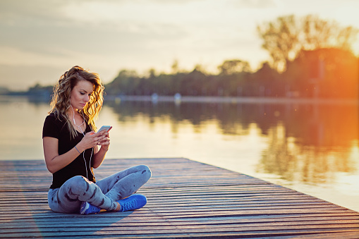 Beautiful girl is texting and listening the music at the front of beautiful sunset