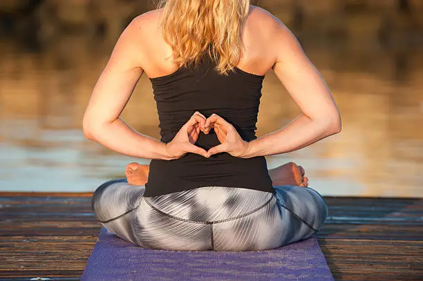 Close view of athletic female seated on dock with hands behind back in heart shape while facing away.