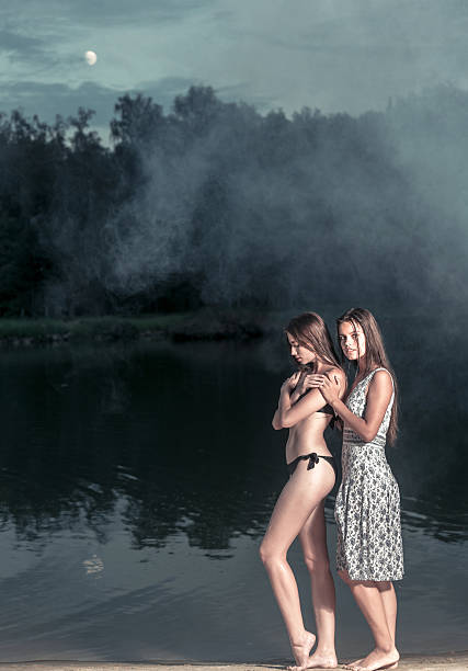 Two beautiful dark-haired girls teenagers are posing together at the forest lake. The girl in bikini is standing with arms folded. The girl in sundress is hugging her friend from the back. She is looking at the camera. Shooting at a mist in warm summer evening outdoors