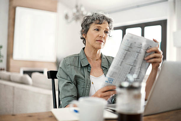 Staying abreast of the latest news Shot of a mature woman reading the newspaper while working on her laptop at home reading newspaper stock pictures, royalty-free photos & images