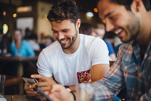 Two male friends talking and using smartphones in a cafe.