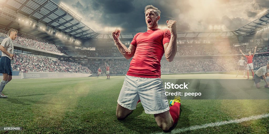 Soccer Player Kneeling on Pitch With Clenched Fists in Celebration A professional male soccer player dressed in red shirt, socks and white shorts kneeling with his arms up and fists clenched, shouting in celebration having just scored a goal. The footballer is on an outdoor pitch, with other teamates and rival players in a generic football soccer stadium full of spectators. Soccer Player Stock Photo