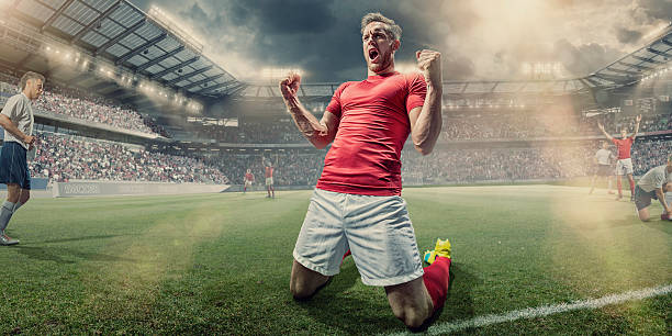 Soccer Player Kneeling on Pitch With Clenched Fists in Celebration A professional male soccer player dressed in red shirt, socks and white shorts kneeling with his arms up and fists clenched, shouting in celebration having just scored a goal. The footballer is on an outdoor pitch, with other teamates and rival players in a generic football soccer stadium full of spectators. soccer player photos stock pictures, royalty-free photos & images