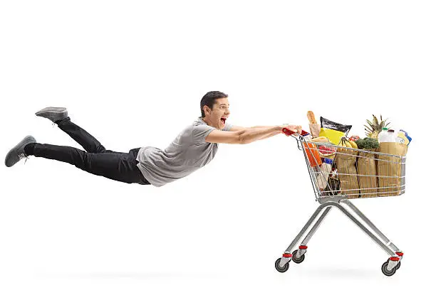 Young man being pulled by a shopping cart full of groceries isolated on white background