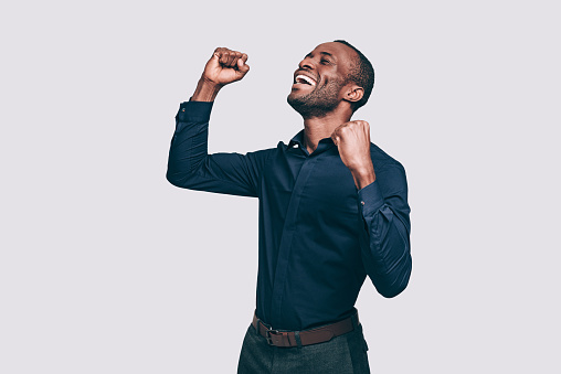 Happy young African man gesturing and keeping eyes closed while standing against grey background