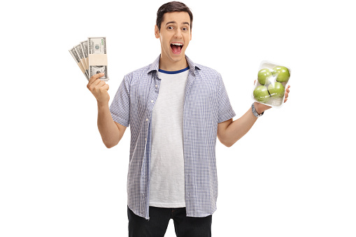 Joyful guy holding bundles of money and a pack of apples isolated on white background