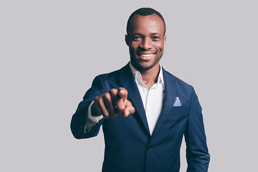 Handsome young African man in smart casual jacket pointing you and smiling while standing against grey background