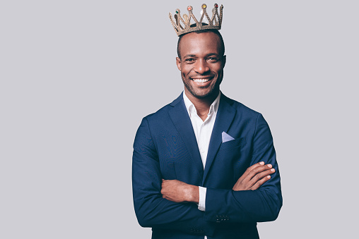 Handsome young African man in crown and smart casual jacket looking at camera and smiling standing against grey background