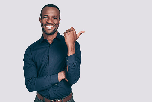 Handsome young African man pointing away and smiling to you while standing against grey background