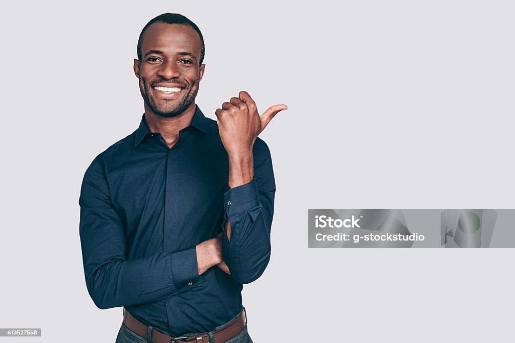 Look over there! Handsome young African man pointing away and smiling to you while standing against grey background Men Stock Photo