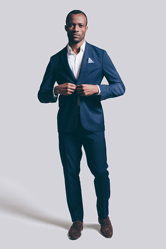 Full length of handsome young African man in full suit buttoning his jacket and looking at camera while standing against grey background