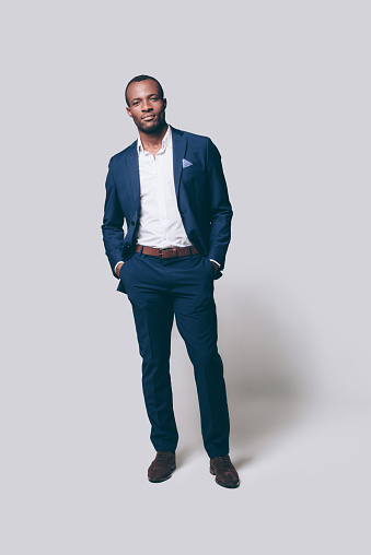 Full length of handsome young African man in smart casual jacket holding hands in pockets and looking at camera while standing against grey background