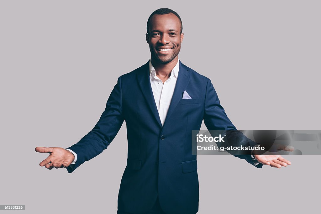 You are welcome! Handsome young African man in smart casual jacket gesturing and smiling while standing against grey background Arms Outstretched Stock Photo