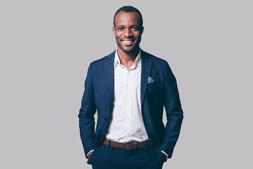 Handsome young African man in smart casual jacket holding hands in pockets and smiling while standing against grey background