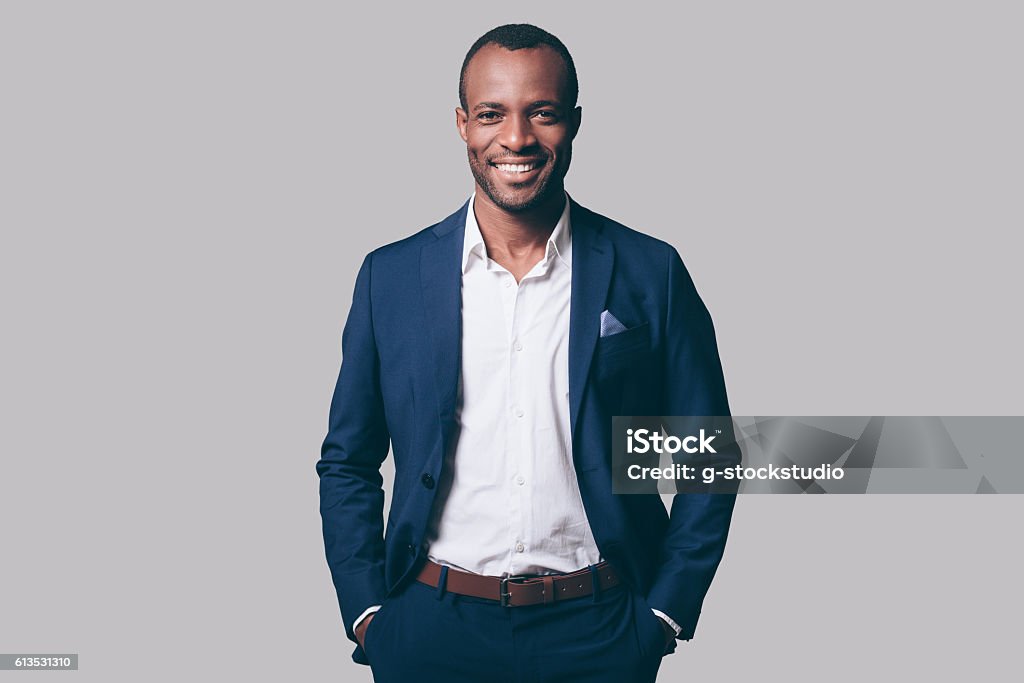 Young and handsome. Handsome young African man in smart casual jacket holding hands in pockets and smiling while standing against grey background Men Stock Photo