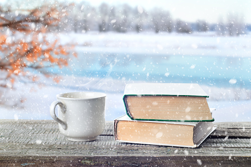 Cup hot coffee or tea, cocoa, chocolate and book outdoors on wooden table or bench in snowy weather on winter season background. Return to winter time. Pile of books and cup in nature.