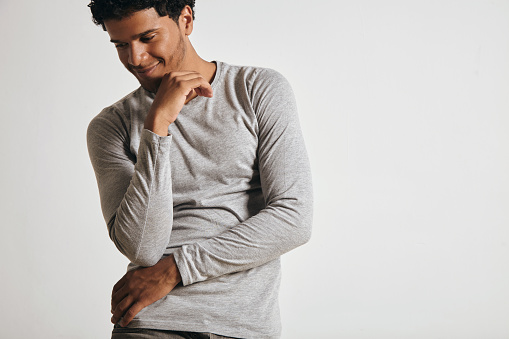 Jouyful smiling young attractive latino guy in blank grey longsleeve looks at side and fposes in front of white wall