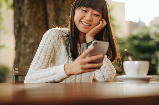 Happy young woman sitting at outdoor cafe and using her cellphone. Female model reading text message on smart phone at coffee shop  and smiling.