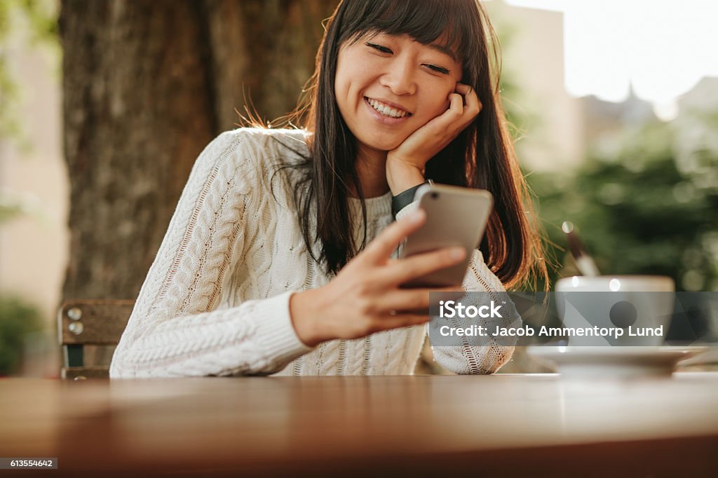 Happy young woman using her cellphone at cafe Happy young woman sitting at outdoor cafe and using her cellphone. Female model reading text message on smart phone at coffee shop  and smiling. Outdoors Stock Photo