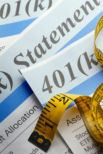 A yellow tape measure sits curled up on top of a 401k account statement. The image conveys the concept of measuring the performance of one's retirement account performance.