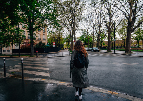 Back side view of young french woman with back pack walking on street of Paris, France in cloudy and rainy day