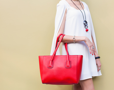 Fashionable beautiful big red handbag on the arm of the girl in a fashionable white dress, posing near the wall on a warm summer night. Warm color. Close up. Part of body.