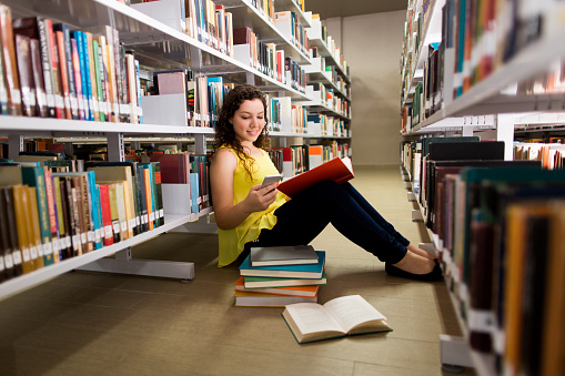 A latin female student reading a book on the floor of a library and being distracted with the mobile phone in a horizontal full length shot indoors.