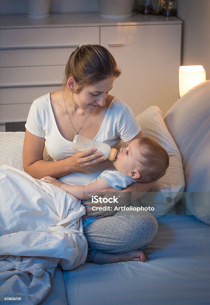 Young mother giving bottle with milk to her baby Portrait of young caring mother giving bottle with milk to her baby in bed at night. Parenting concept Baby - Human Age Stock Photo