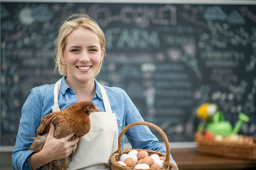 A female entrepreneur and farmer is selling eggs and produce at the farmers market. She is holding a chicken and a basket of eggs.
