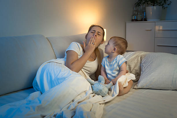 Baby boy sitting on bed and looking at yawning mother 