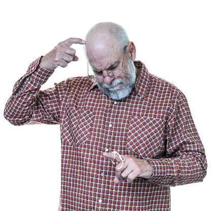 A frustrated, forgetful senior adult man is embarrassed and befuddled because he can't remember why he tied the knot in that string around his index finger as a reminder. He is scratching his head with his other hand index finger as he ponders which forgotten chore needs his attention. He is a grey-bearded, balding, eyeglass wearing, mid-60's year old baby boomer dressed in a plaid long sleeve button-up shirt with a collar. For concepts including general problems related to human aging, busy lifestyle distractions, carelessness, attention deficit disorder, dementia, Alzheimer's Disease, and other mental illness and/or senior citizen healthcare and medicine issues and concerns. White background with copy space. Canon 5D Mark III.